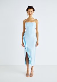 Robe en satin bleu clair avec un décolleté carré, des bretelles fines, un détail de volants asymétriques sur un côté et une fente haute sur la cuisse. Les accessoires incluent des talons strappy.