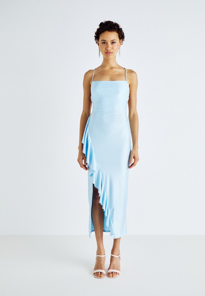 Robe en satin bleu clair avec un décolleté carré, des bretelles fines, un détail de volants asymétriques sur un côté et une fente haute sur la cuisse. Les accessoires incluent des talons strappy.