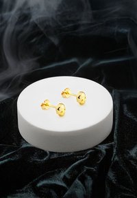 Guldörhängen med en lystrande yta, med rundad design och små rosettaccents, uppställda på en vit cirkulär pedestal mot en mörk bakgrund.