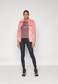 Rosa plys fleece-jakke med høy krage, brukt over en mørk lilla grafisk t-skjorte. Svart, tettsittende bukser med kneputer og turstøvler.