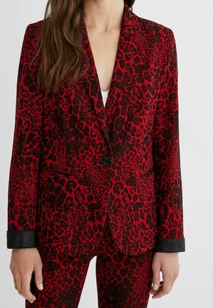 Woman wearing a red and black leopard print blazer with black cuffs, buttoned at the waist, paired with matching pants.