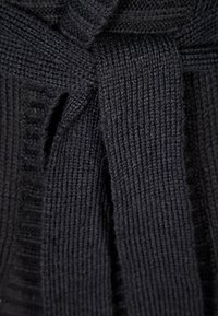 Czarna dzianina o grubym prążkowanym splocie związana w węzeł lub kokardę, ukazująca szczegółowe ściegi i miękki materiał.