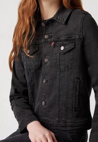 Chaqueta de mezclilla negra con un diseño clásico, que cuenta con dos bolsillos en el pecho, cierre frontal con botones y detalles de metal. Tela texturada.
