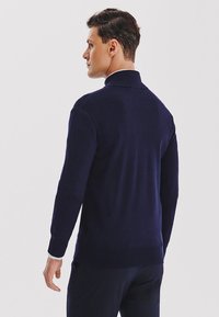 Granatowy sweter z dzianiny, z kołnierzem, ściągaczami na rękawach, gładką fakturą i dopasowanym krojem. Pokazany od tyłu.