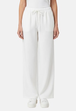 Donna che indossa pantaloni bianchi larghi con coulisse e sneaker bianche, in piedi davanti a uno sfondo chiaro e uniforme.