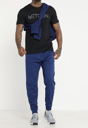 Svart t-shirt med logo, marinblå joggers och grå träningsskor. Sweatshirt hängd över axlarna. Mjukt tyg, avslappnad passform.