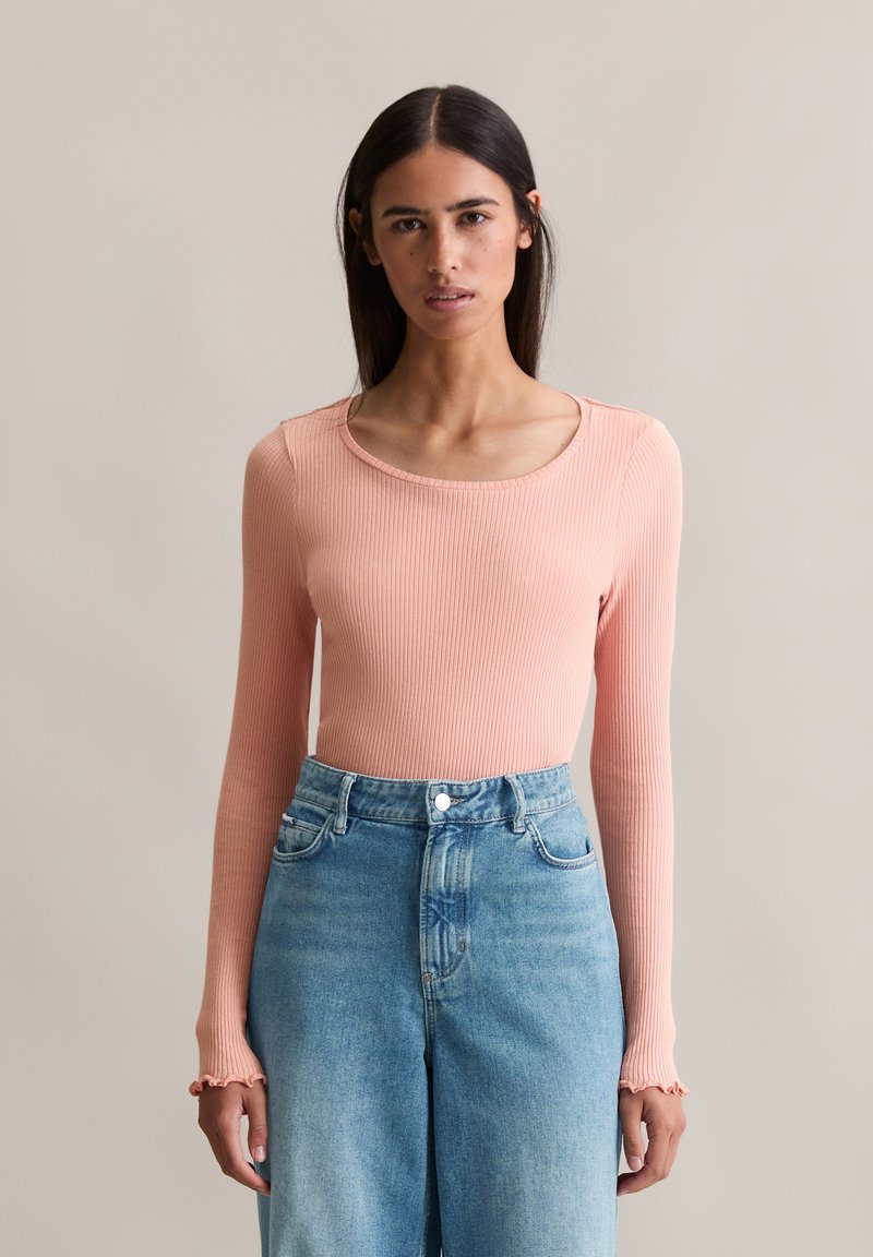 Haut à manches longues en rib, de couleur pêche douce, avec un décolleté arrondi et des poignets à volants, associé à un jean en denim bleu clair taille haute.