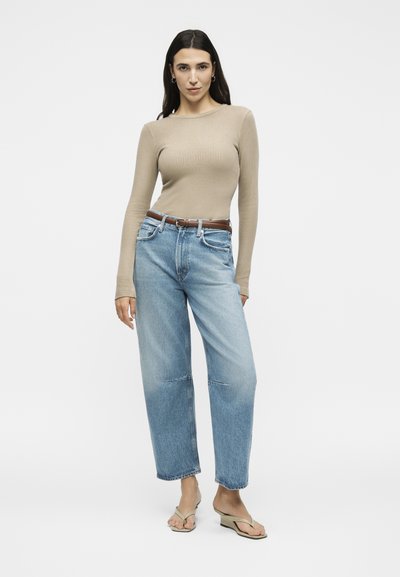 Γυναίκα που στέκεται, φοράει μπεζ ριμπ μακρυμάνικη μπλούζα, ανοιχτό μπλε ψηλόμεσα τζιν με καφέ ζώνη και μπεζ πλατφόρμες σανδάλια.