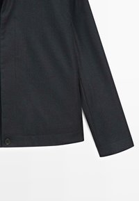 Ciemnoszary żakiet z gładką fakturą, z pojedynczym zapięciem na guzik i konstrukcyjnymi ramionami. Dopasowany krój z długimi rękawami.