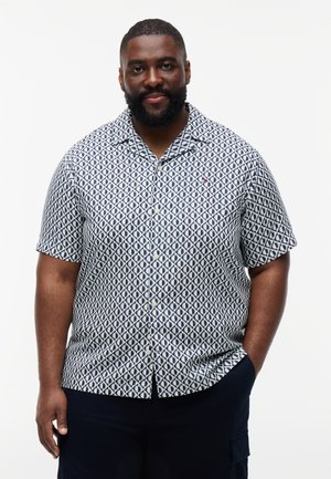 Mężczyzna z brodą, ubrany w krótki rękaw, wzorzystą koszulę na guziki i ciemne spodnie, stojący z jedną ręką w kieszeni na białym tle.