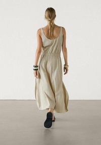 Robe maxi beige sans manches avec une taille élastique, un tissu fluide et un dos échancré. Portée avec des chaussures noires sans lacets et des bracelets.
