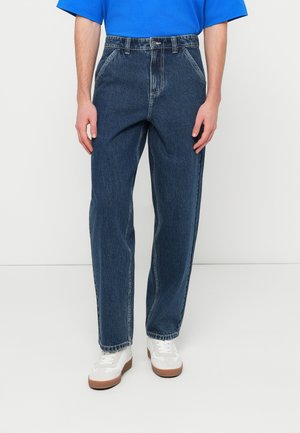 Άνδρας που φοράει άνετο, διχρωματικό τζιν σκούρου μπλε και λευκά αθλητικά παπούτσια, στέκεται μπροστά σε ένα απλό φωτεινό φόντο.