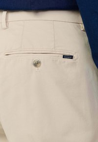 Pantalon beige sur mesure avec une poche arrière boutonnée et une petite étiquette de la marque Hackett sur le bord de la poche, porté avec un haut bleu foncé.