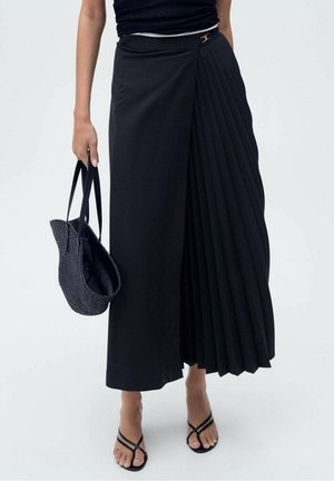 Gonna nera con design asimmetrico, caratterizzata da un frontalino liscio e un lato plissettato. Abbinata a una borsa intrecciata e sandali con cinturini.