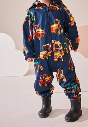 Enfant portant une combinaison à capuche bleu marine avec des imprimés colorés de véhicules de construction et des bottes en caoutchouc noires, debout sur un sol clair.