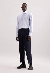 Camicia bianca a quadretti con colletto appuntito, abbinata a pantaloni blu navy e scarpe di pelle nere, in piedi davanti a uno sfondo neutro.