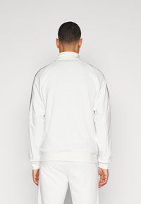 Pullover in fleece bianco con colletto alto, maniche lunghe e tasche laterali. Presenta una texture liscia e strisce sottili di accento sulle maniche.