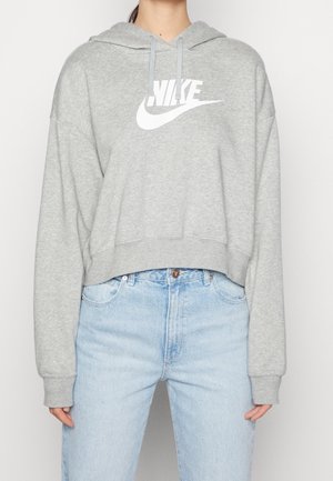 Person iført en lysegrå Nike hættetrøje med hvidt logo og lyseblå højtaljede jeans mod en ensfarvet baggrund.