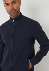 Maglione zip-up blu navy con colletto alto e polsini a costine. Presenta tasche laterali e un tessuto a maglia dalla superficie liscia.