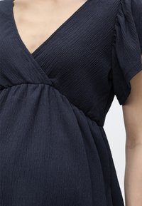 Γυναίκα που φοράει φόρεμα από σκούρο υφασμάτινο ύφασμα με V-λαιμόκοψη και κοντά, φουσκωτά μανίκια, εμφανίζοντας τον άνω κορμό και τη μέση.