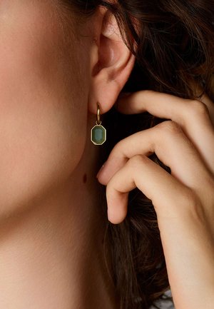 Gouden hoepeloorbel met een octagonale groene steen. De oorbel is bevestigd aan de oorschelp, met een hand die voorzichtig het haar nabij aanraakt.
