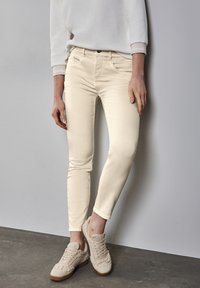 Vaqueros skinny beige con una textura suave, cintura media y bolsillos inclinados, combinados con zapatillas beige texturizadas sobre una superficie gris.