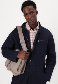 Σκούρο μπλε μπουφάν εργασίας με μπροστινό φερμουάρ, πλάγιες τσέπες και μια καρό ανοιχτού χρώματος μπλούζα από κάτω. Γκρίζα τσάντα ώμου από ύφασμα με φερμουάρ.