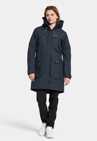 Marineblauwe waterdichte parka met capuchon, twee voorzakken en trekkoorddetails. Heeft een licht uitlopende vorm en een gladde textuur.