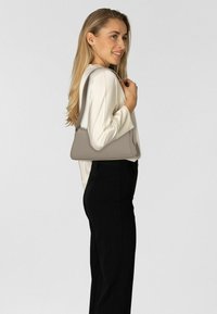 Mujer con cabello largo y rubio, vistiendo una blusa crema y pantalones negros, sosteniendo un bolso de hombro color topo sobre un fondo gris liso.