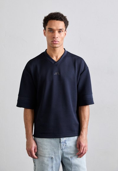 Navy blue short-sleeve v-neck shirt with a textured surface and logo detail at chest. Paired with light blue denim pants.