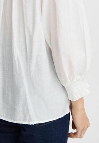 Vit blus med texturerad tyg, elastiska puffärmar och en avslappnad passform. Bärs med mörka jeans som visar sid sömn detaljer.