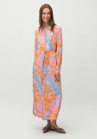 Blumenmuster, langärmliges Kleid in Blau, Pink und Orange. Mit Knopfleiste, Taillengürtel und weichem Stoff; getragen mit flachen Schuhen.