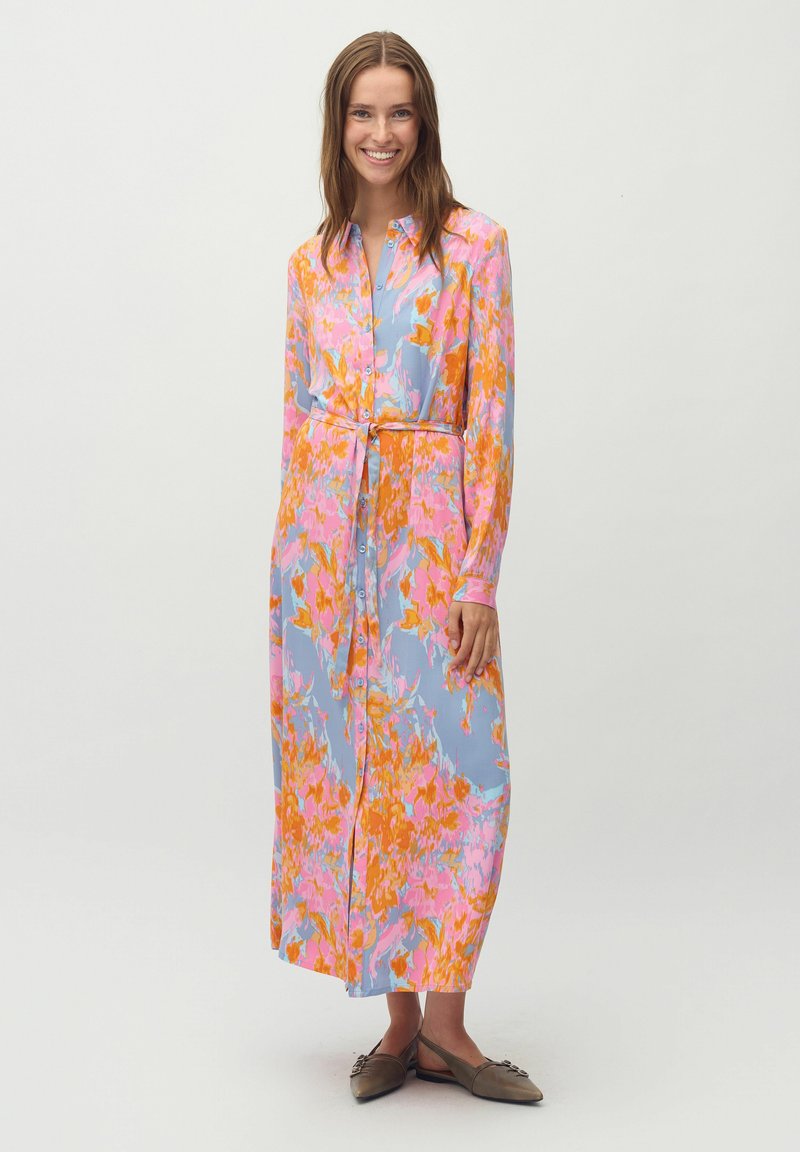Blumenmuster, langärmliges Kleid in Blau, Pink und Orange. Mit Knopfleiste, Taillengürtel und weichem Stoff; getragen mit flachen Schuhen.