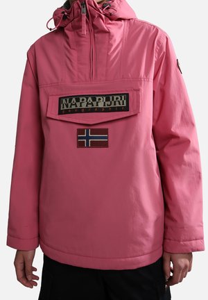 Roze capuchonjas met voorrits, grote Napapijri logo patch en Noorse vlag embleem, gedragen door een persoon met ontspannen handen langs het lichaam.