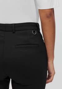 Zwarte high-waisted broek met een gladde textuur, voorzien van riemlussen, een cirkelvormig hardware detail en een achterzak.