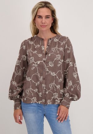 Vrouw in een bruine bloemetjes blouse met lange pofmouwen en lichtblauwe jeans, staand tegen een effen achtergrond.