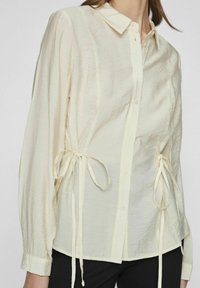 Chemise claire boutonnée avec une coupe ample, dotée de longues manches, de liens à la taille et d'une surface en tissu texturé.