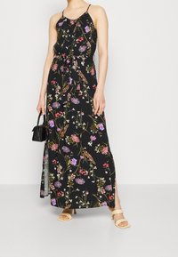 Robe maxi noire à fleurs avec des bretelles fines, présentant des motifs de fleurs colorées et une taille nouée. Les détails incluent des fentes latérales et un tissu léger.
