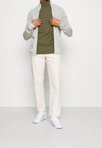 Suéter gris claro con cremallera sobre un suéter de cuello alto verde oliva, combinado con pantalones de color crema y zapatillas blancas. Texturas suaves, diseño casual.