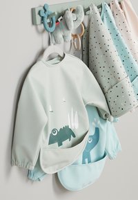 Lichtgroene waterdichte schort met een donkergroene dinosaurusafbeelding; beige, muntgroene en patroonbedrukte tassen hangen naast elkaar aan een haak.