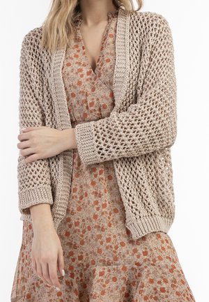 Beige gebreide cardigan met een open weefsel, met lange mouwen en geribbelde manchetten, gedragen over een bloemenjurk in warme kleuren.