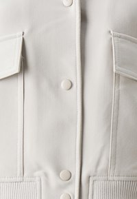 Chemise en tissu blanc avec une finition texturée, dotée de deux poches à l'avant, de boutons et d'accents côtelés le long de l'ourlet et des poignets.