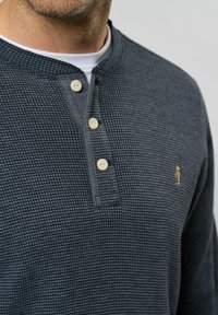 Tamno plava Henley majica s teksturom vafla, okruglim izrezom, tri bež dugmeta i malim zlatnim logom na lijevoj strani prsnog koša.