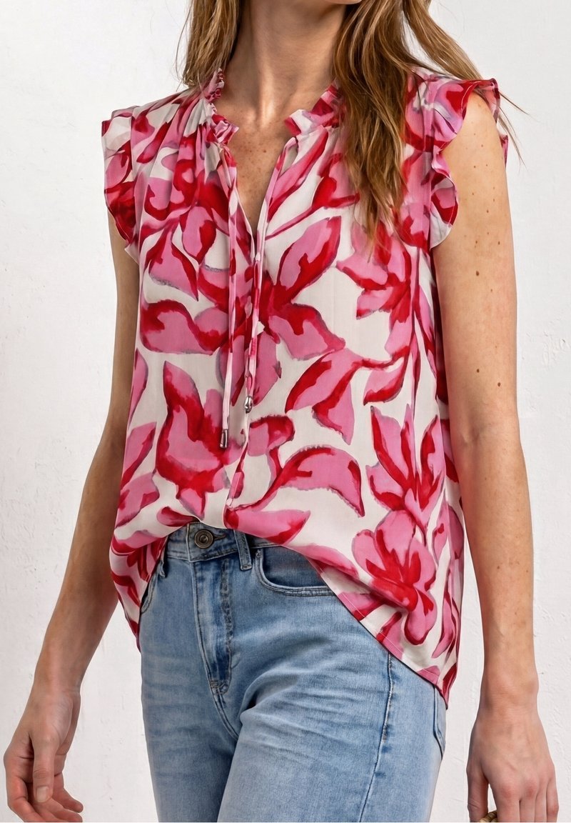 Femme portant un chemisier sans manches à motif floral rose et rouge rentré dans un jean bleu clair sur un fond clair uni.