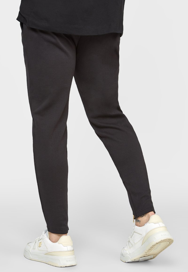 Mustat kapealahkeiset collegehousut, valmistettu puuvillasekoitteesta, joissa on sileä pinta ja nilkassa vetoketjuyksityiskohta, yhdistetty valkoisiin lenkkareihin.