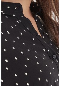 Camicia nera con bottoni a pressione, decorata con pois bianchi, colletto e tessuto testurizzato con finitura liscia. Due bottoni neri visibili.