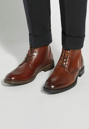 Brown leather ankle boots feature a smooth texture, lace-up front, and a low heel, paired with dark cuffed trousers.
