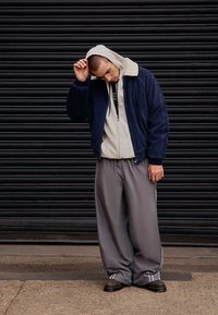 Μπουφάν bomber από ναυτικό κορδόνι με κρεμ γιακά shearling, φορεμένο πάνω από γκρι ριγωτό φούτερ με κουκούλα και άνετο γκρι παντελόνι με ρίγες στο πλάι. Μαύρα μποτάκια.