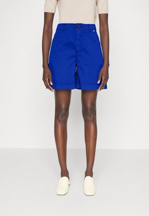 Personne portant un short bleu vif cintré, un haut beige à manches courtes et des chaussures blanches à enfiler, debout devant un fond clair uni.