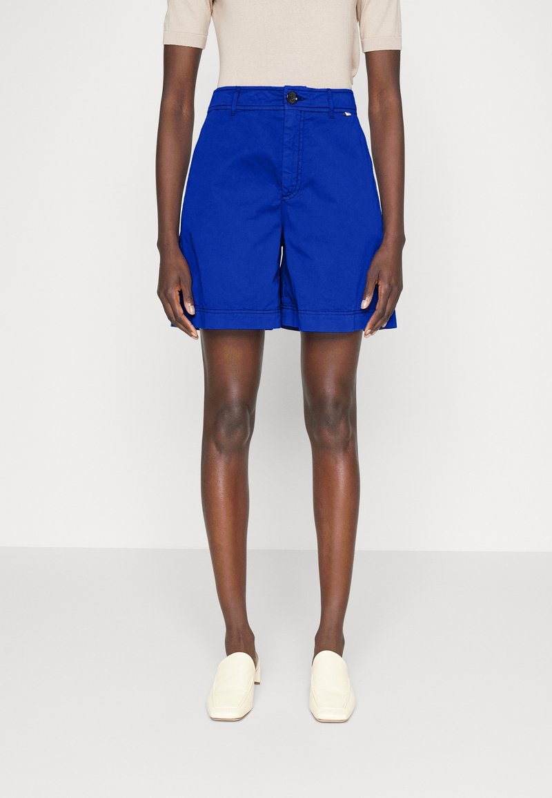 Personne portant un short bleu vif cintré, un haut beige à manches courtes et des chaussures blanches à enfiler, debout devant un fond clair uni.